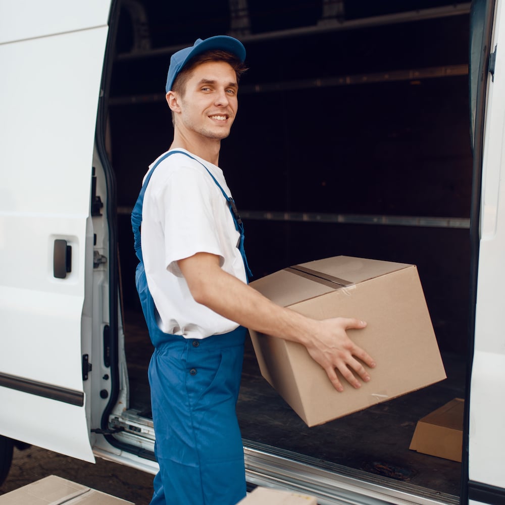 Deliveryman in uniform unloads the car with parcels, delivery service. Man standing at cardboard packages in vehicle, male deliver, courier or shipping job